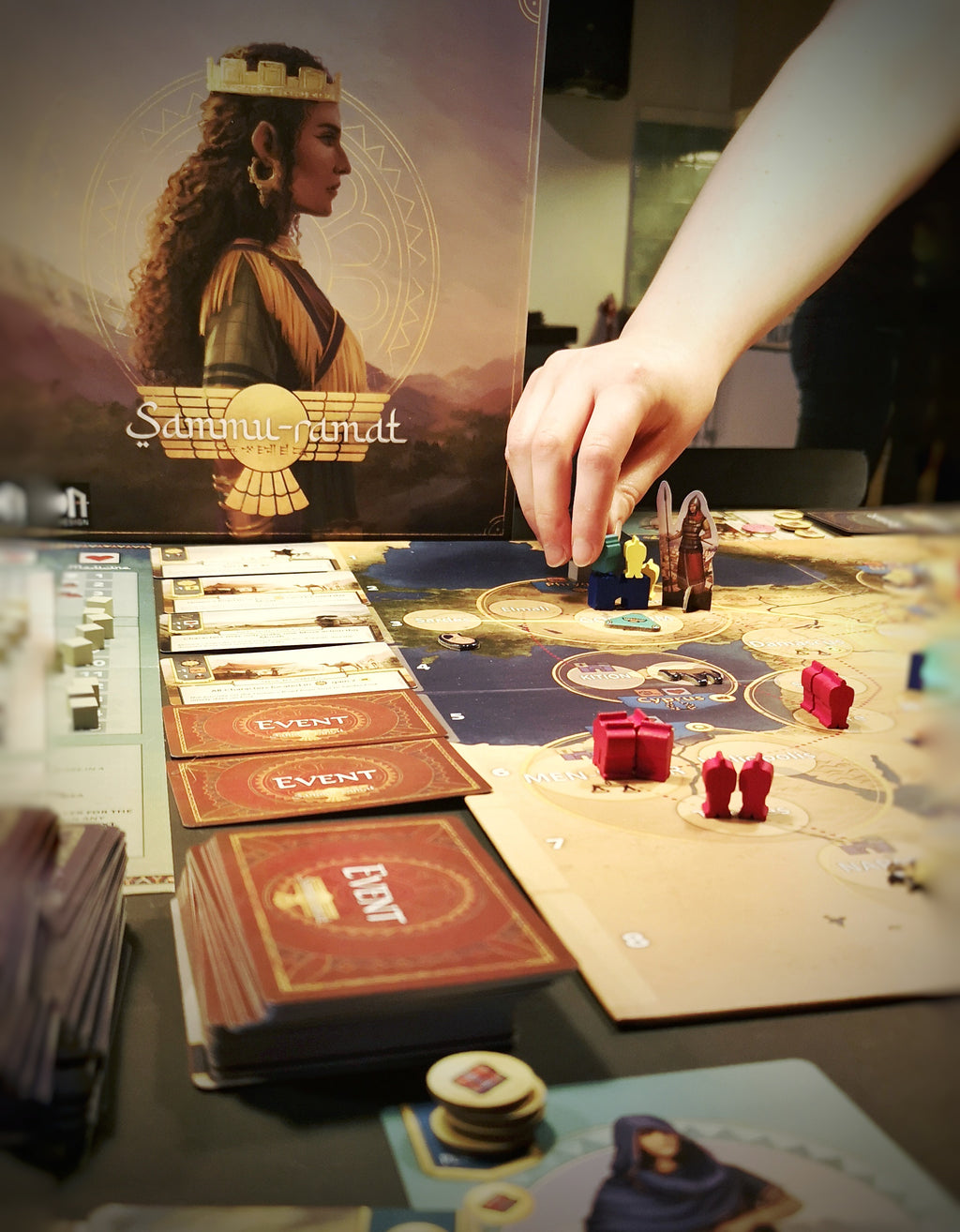 A hand reaches down to move game components on the board. Piles of cards lie beside the board, and the box is standing in the background.